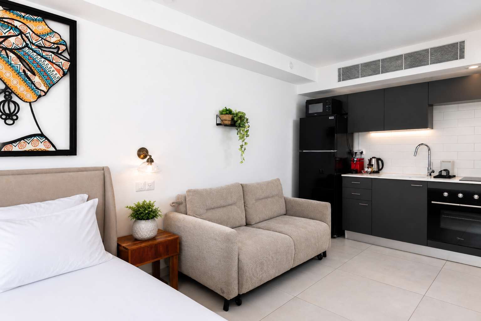 Una camera d'albergo moderna con un letto, una poltrona, un tavolino con una pianta, e un angolo cottura dotato di microonde, frigorifero e lavello.
