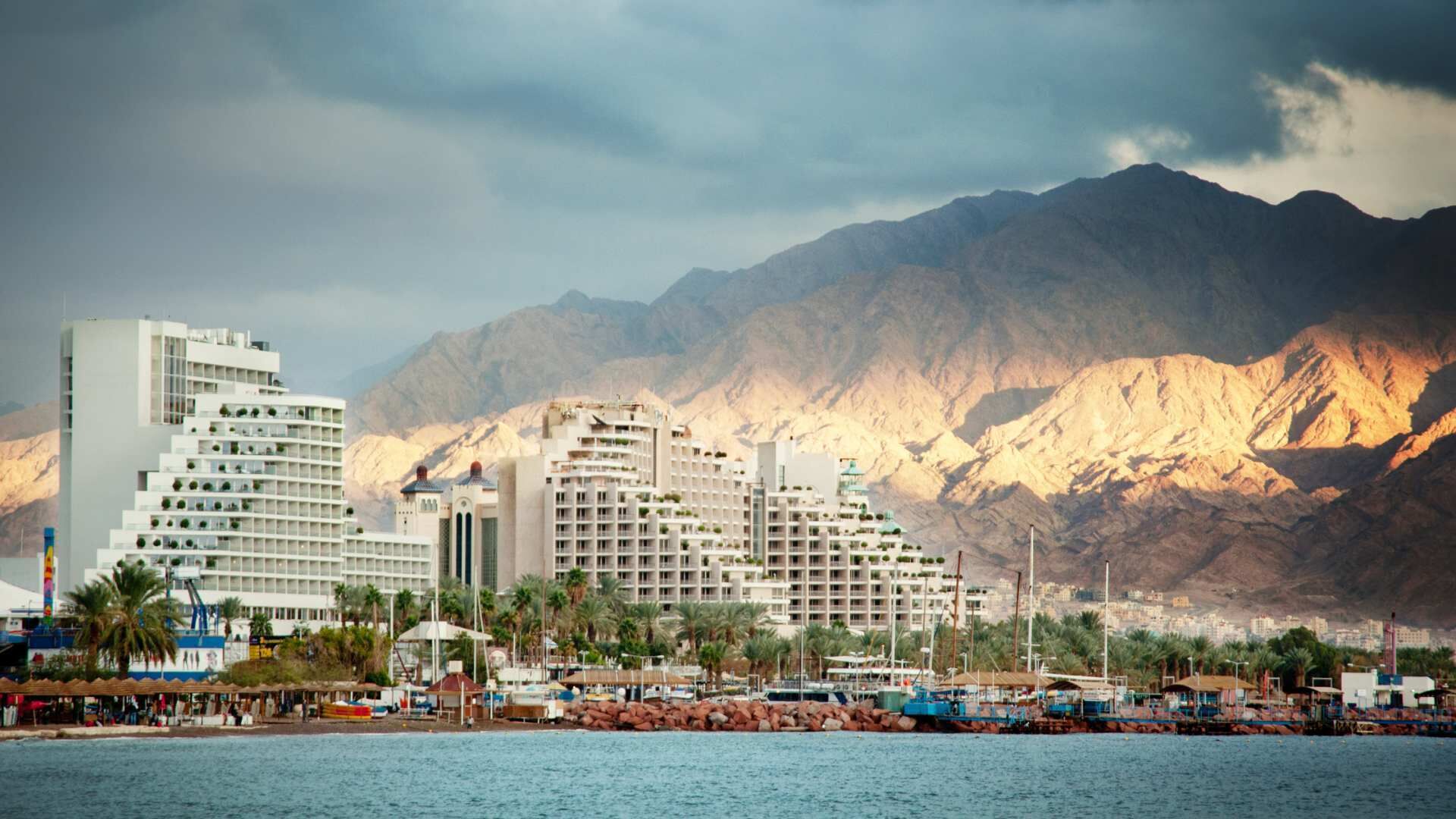 A coastal hotel with multiple buildings, a marina with boats, and a mountainous backdrop.