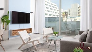 Ein modernes Wohnzimmer mit einem an der Wand befestigten Flachbildfernseher, einer Couch, zwei gemusterten Sesseln, einem kleinen Tisch mit einer Pflanze und einem Balkon mit Glastrennwand.
