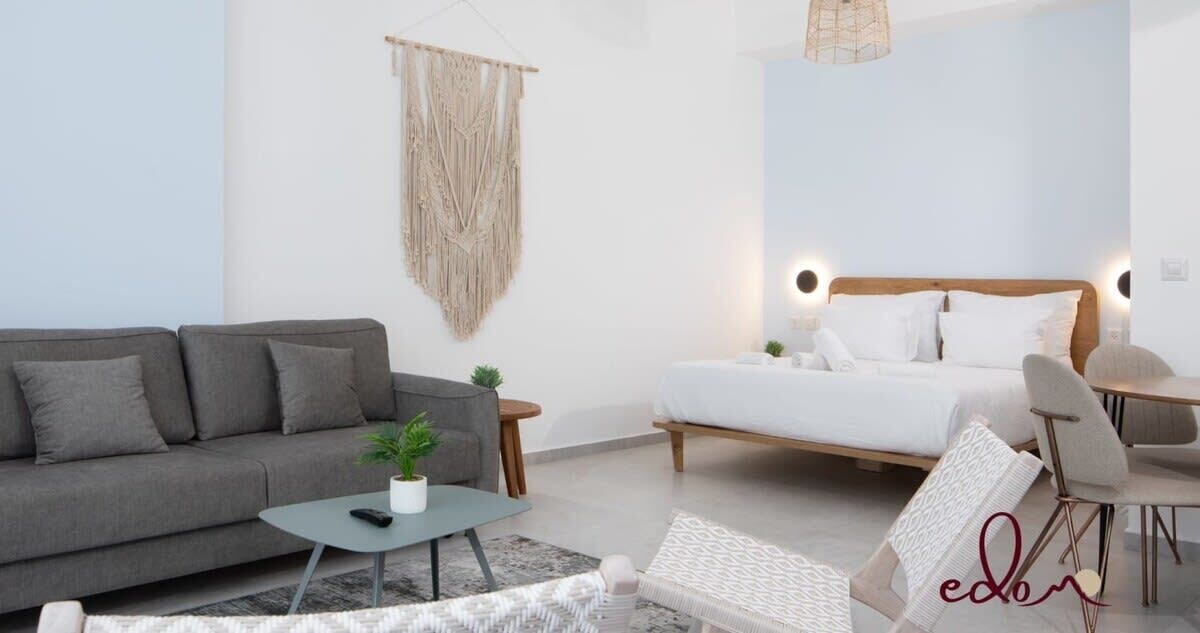 A modern hotel room with a grey sofa, a small table with a plant, a bed with white bedding, and a hanging macrame wall decoration.