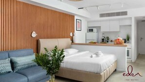 Una habitación de hotel moderna con cama, sofá, paneles de pared de madera, zona de cocina y una planta pequeña.