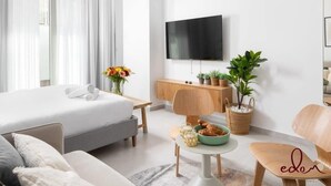 A modern hotel room with a bed, a TV, a wooden desk, a small table with a bowl of snacks, and a potted plant. - דירת גן מפנקת בסיסייד 100 מטר מהים - By Edom (Eilat)