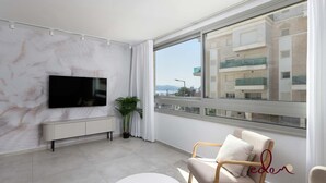 A modern living room with a large window, a flat-screen TV, a white radiator, and a potted plant. - סוויטה מעוצבת במתחם סיי סייד - By Edomlux (Eilat)