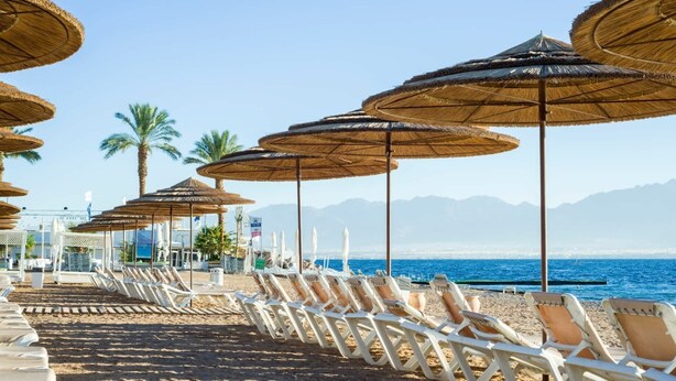 Een strand met rijen ligbedden en rieten parasols, palmbomen en een uitzicht op zee en bergen op de achtergrond.