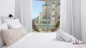 Ett hotellrum med en säng, vita gardiner, utsikt över en byggnad och en stol vid fönstret.