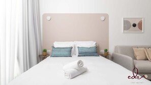 A neatly made bed with two pillows, a folded towel, and a headboard with two circular cutouts. There is a small table with a plant and a framed picture on the wall. - דירת גן קלאסית בסי סייד 50 מטר מהים - By Edom (Eilat)