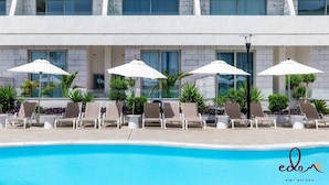 Área de piscina com espreguiçadeiras e guarda-sóis, piscina azul cristalina e fachada de edifício moderna.
