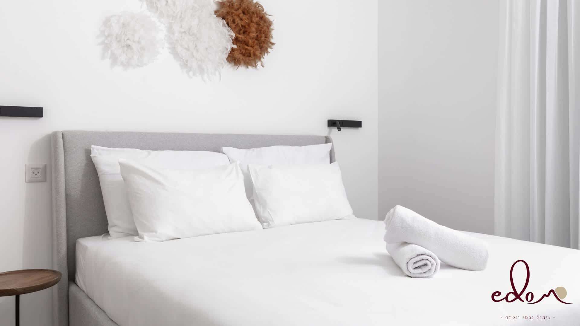Una cama bien hecha con ropa de cama blanca, cabecera gris y una toalla blanca enrollada al lado.