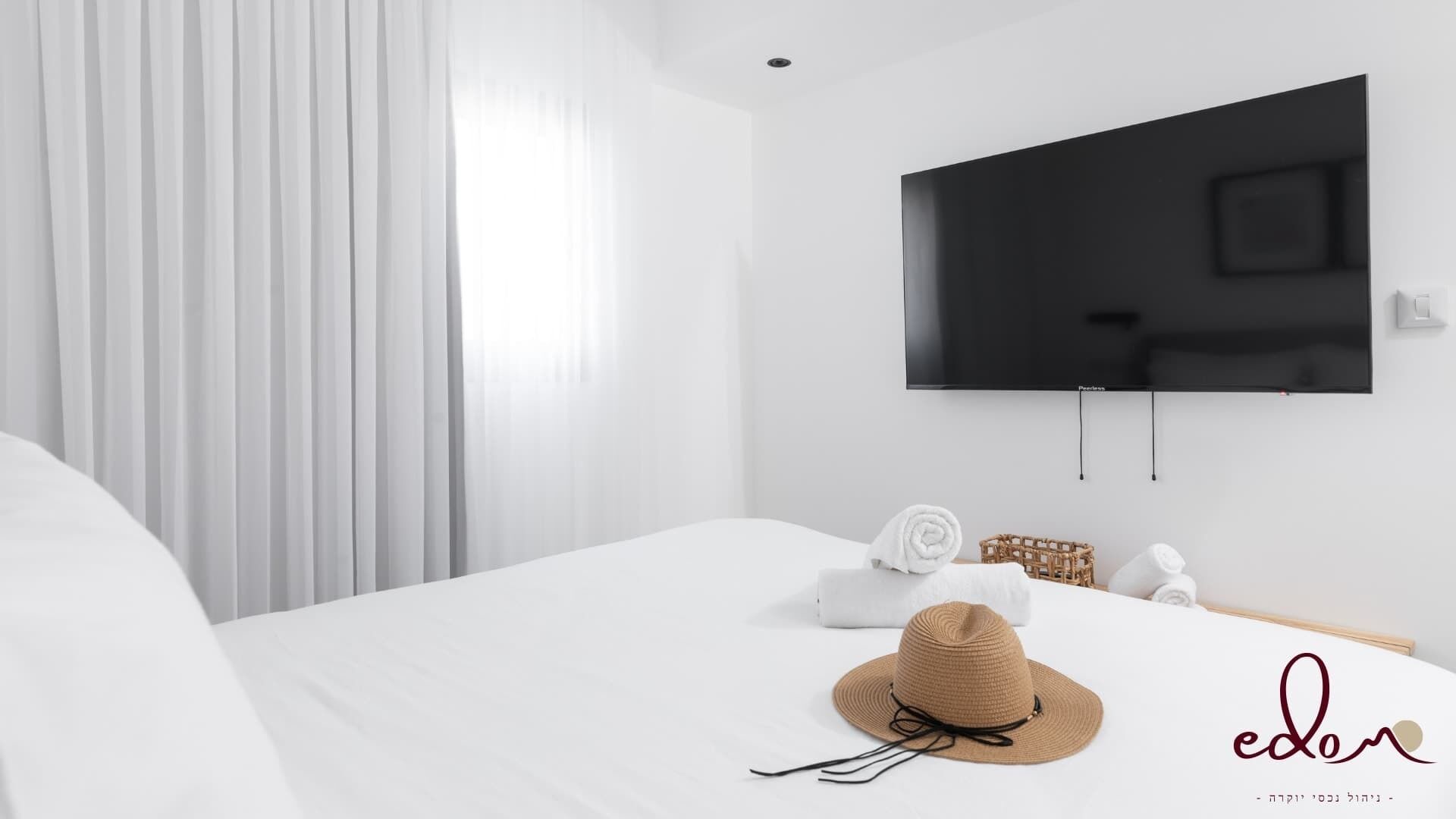 Quarto de hotel moderno com uma cama grande, uma TV de tela plana na parede e uma janela com cortinas brancas.