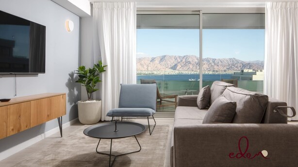A modern living room with a flat-screen TV, a grey sofa, a blue armchair, a round coffee table, and a large window offering a mountain view. - דירת 2 חדרי שינה עם נוף מדהים לים - By Edom (Eilat)