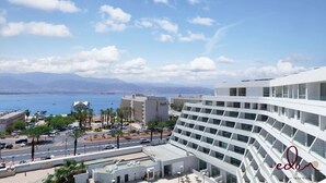 Hotel moderno com vista para o mar e montanhas. Possui amplo estacionamento, palmeiras e céu claro.