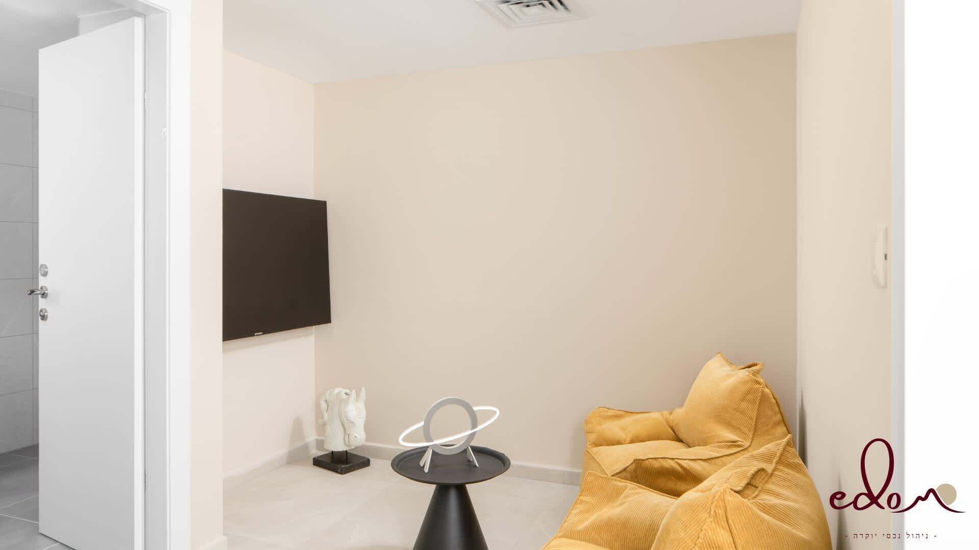 Habitación minimalista con una puerta blanca, un televisor de pantalla plana montado en la pared, una mesita con un objeto decorativo y una silla amarilla.