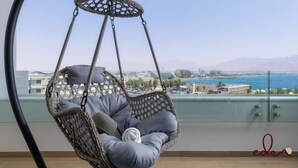 A hanging chair with cushions on a balcony overlooking a cityscape and water. - מיני פנטהאוז 2 חדרי שינה במתחם סי סייד - By Edom (Eilat)