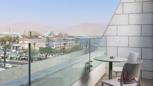 A balcony with a glass railing, a small round table, and two chairs. A cityscape with buildings and a body of water is visible in the background. - דירה בסי סייד עם נוף מדהים לים - By Edom (Eilat)