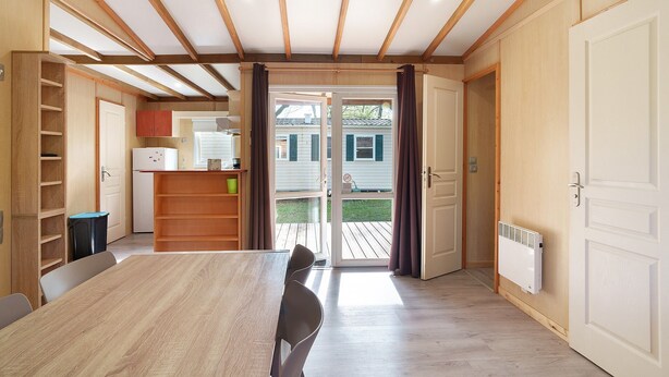 A room with a wooden ceiling, a dining table, chairs, a white radiator, and a sliding glass door leading to an outdoor area. - Chalet “Le Convivial Cottage” with Shared Pool, Wi-Fi, and Air Conditioning (Saint-Martial-de-Nabirat)