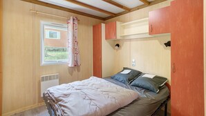 A bedroom with a bed, a window with curtains, a radiator, and wooden ceiling beams. - Chalet “Le Convivial Cottage” with Shared Pool, Wi-Fi, and Air Conditioning (Saint-Martial-de-Nabirat)