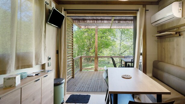 Ein Zimmer mit Holztisch, einer Bank, einem an der Wand befestigten Fernseher und Blick auf Grünflächen.