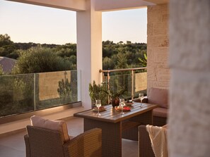 A balcony with wicker furniture, a table set with a glass of wine, and a view of greenery. - Solis Ortus Villa, a Sublime Sanctuary with Pool (Zakynthos)