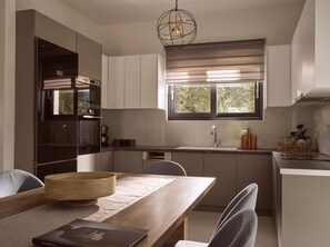 A modern kitchen with a dining area, featuring a built-in oven, a large window with blinds, and a hanging light fixture. - Solis Ortus Villa, a Sublime Sanctuary with Pool (Zakynthos)