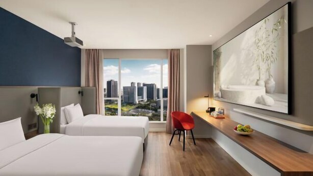 Ein Hotelzimmer mit zwei Betten, einem Schreibtisch mit Stuhl, einem großen Fenster mit Stadtblick und einem aufgehängten Kunstwerk.
