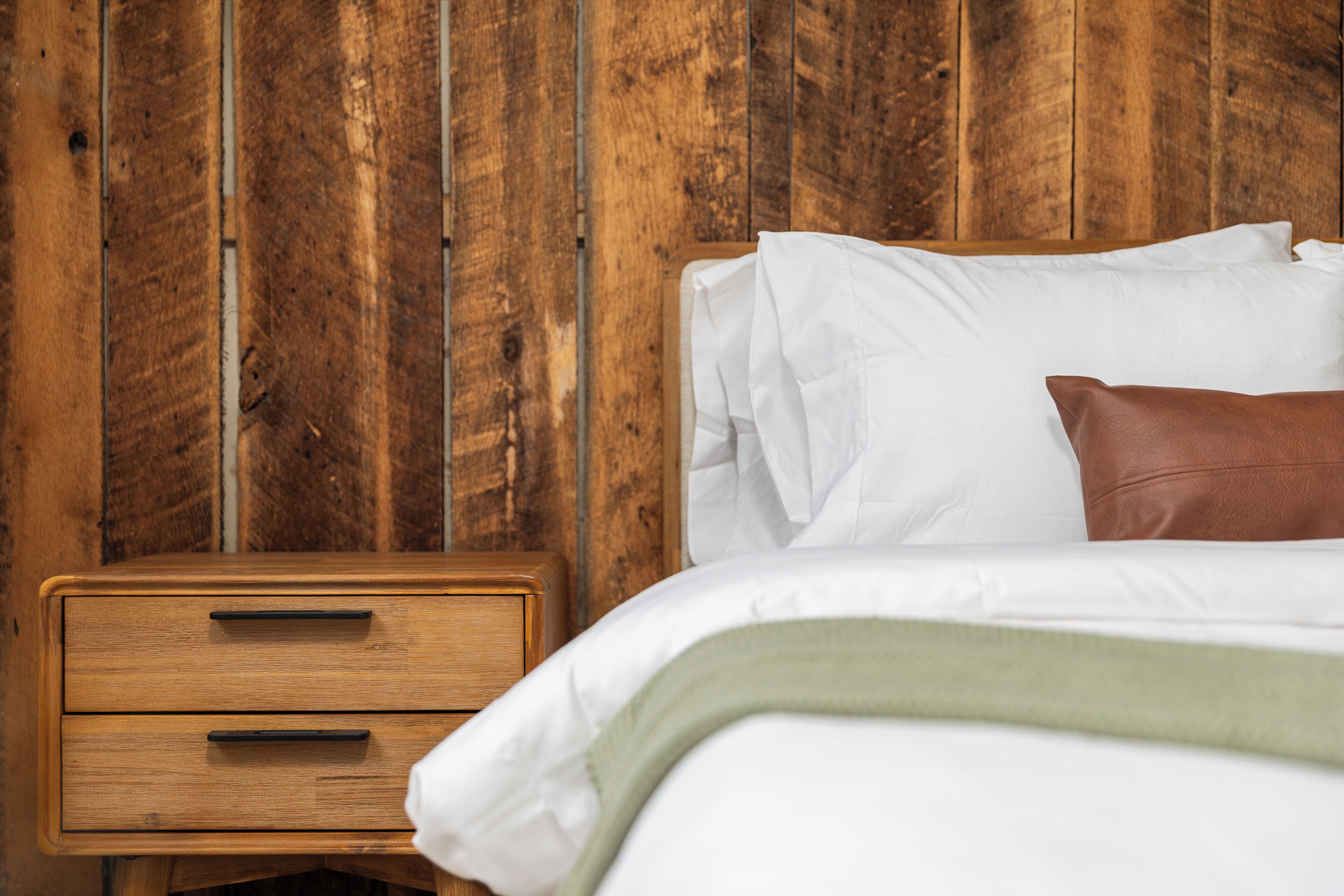Uma mesa de cabeceira de madeira com gavetas, uma cama com roupa de cama branca e um cobertor verde, e uma parede de madeira.