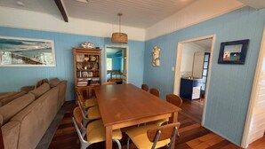 A dining area with a wooden table and chairs, a bookshelf, and a view of a bedroom through an open door. - By The Sea - by Lifestyle. Brunswick Heads (NEW BRIGHTON)