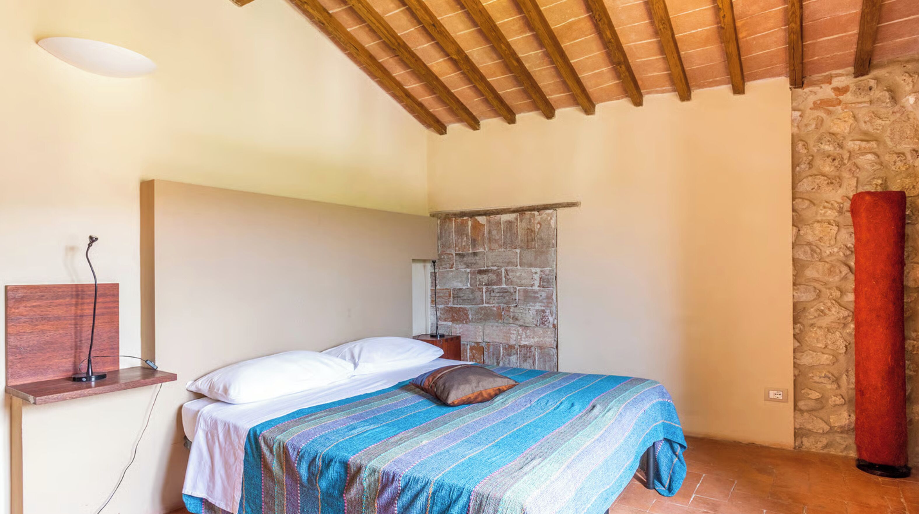 A bedroom with a bed, a nightstand, a stone wall, and a wooden ceiling.