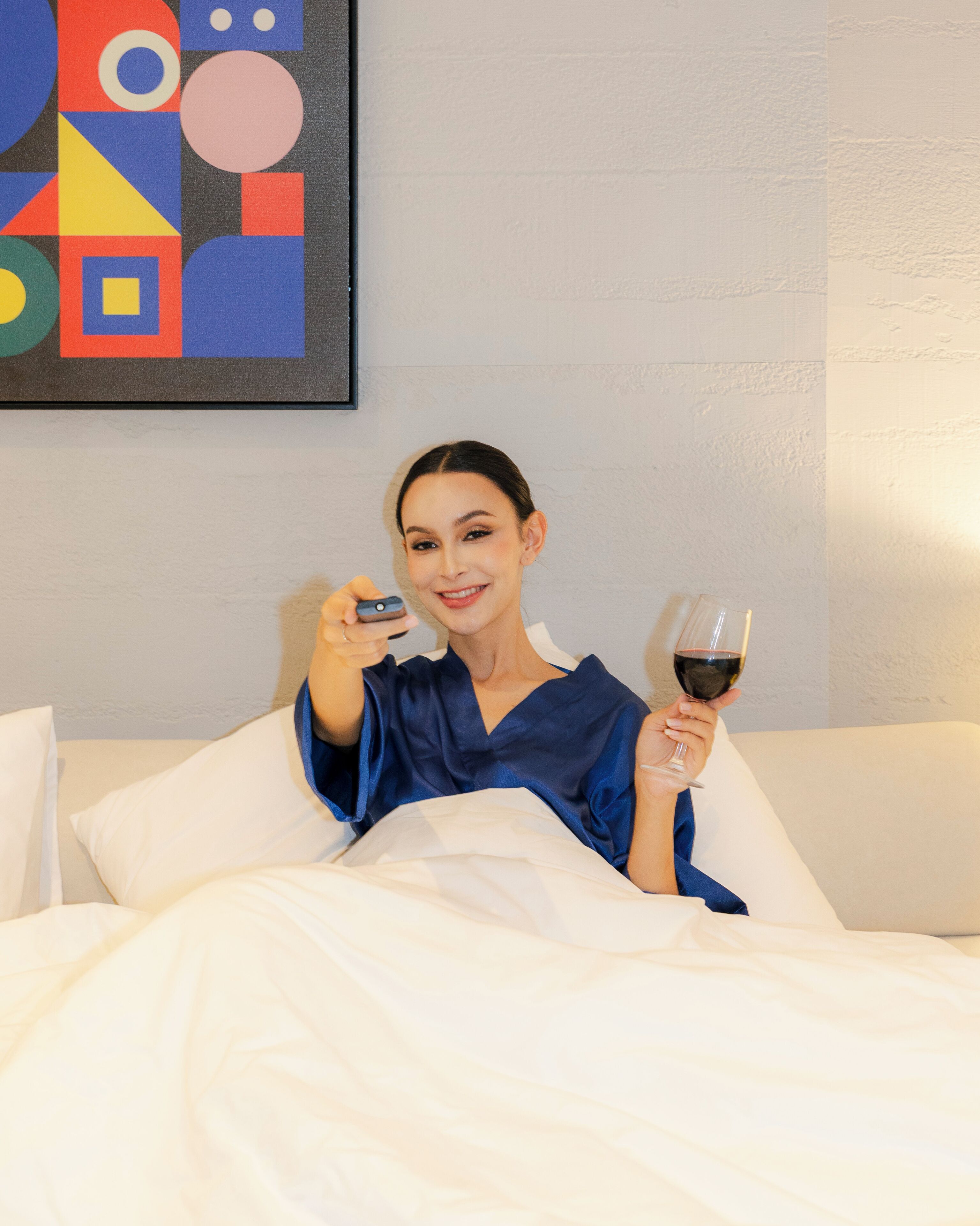 A person in a blue robe sitting on a bed with white bedding, holding a smartphone and a wine glass.