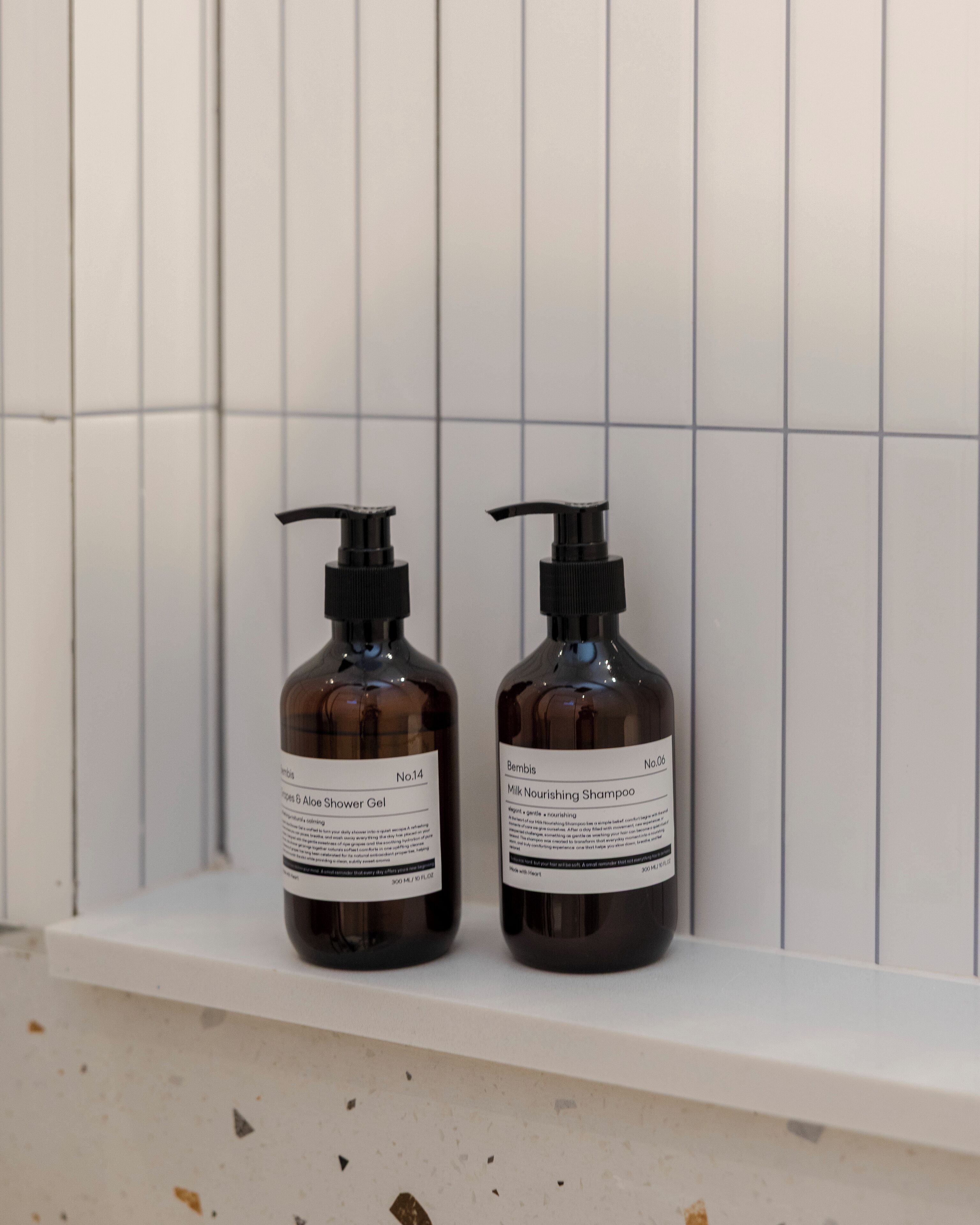 Two bottles of shower gel and shampoo on a white shelf against a tiled wall.