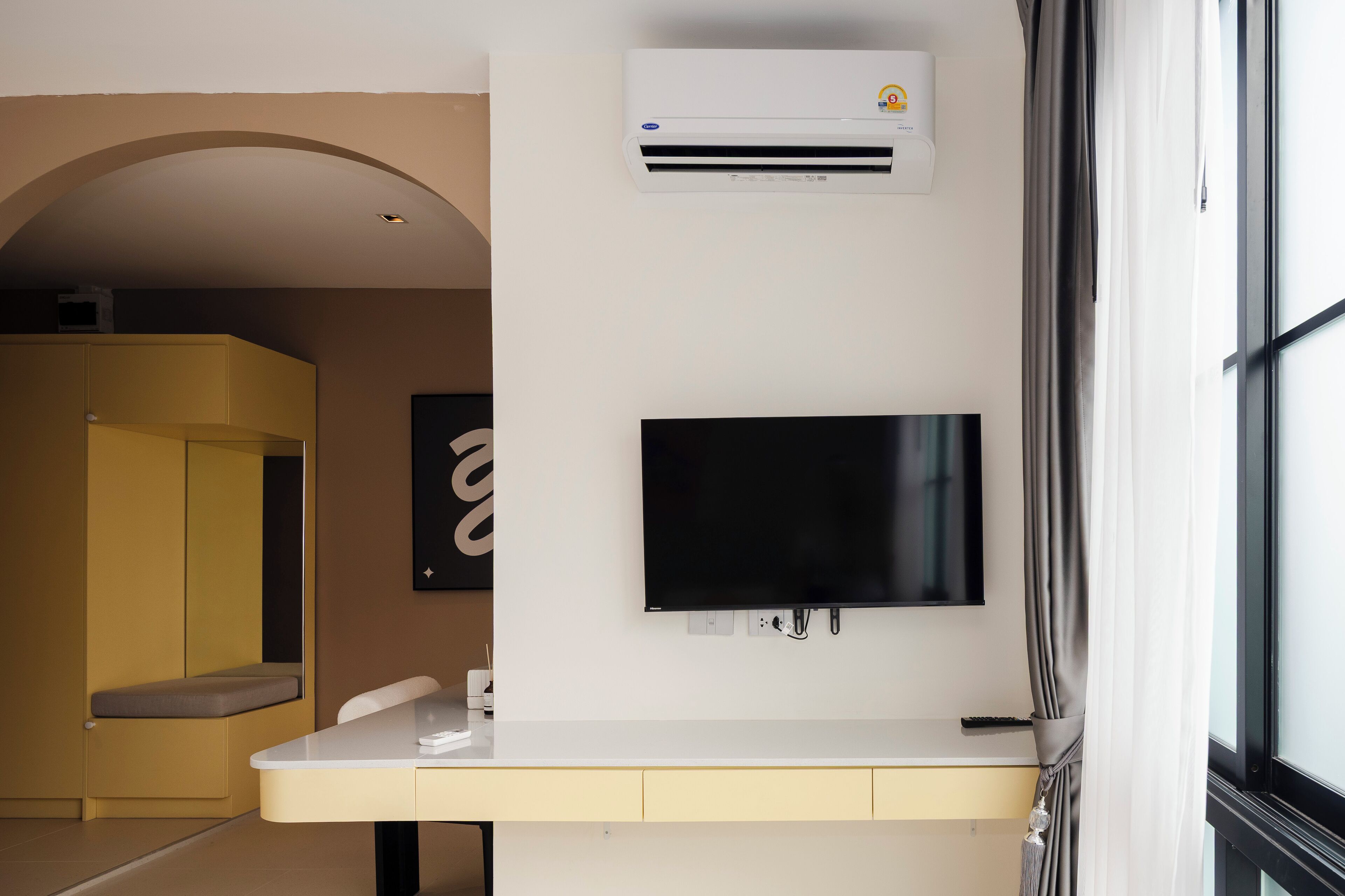 A modern hotel room with a flat-screen TV mounted on the wall, a built-in air conditioning unit, and a window with curtains.