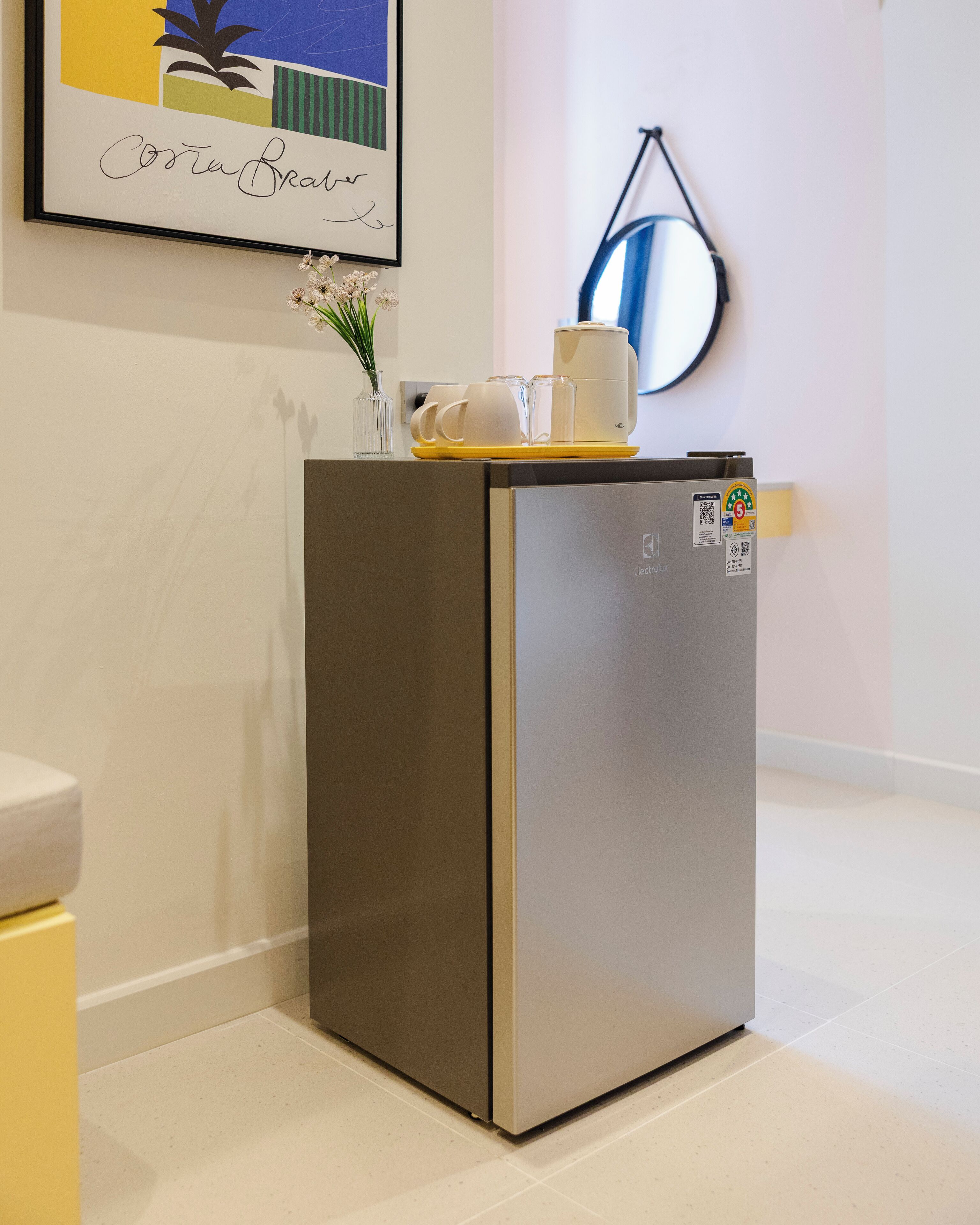A compact, modern refrigerator in a clean, minimalistic room with a framed artwork on the wall and a small vase with flowers on top.