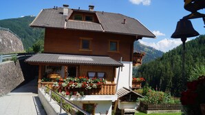 A two-story wooden house with a balcony, a stone wall, and a garden with flowers. - Traditional Apartment "Waldstille" in Idyllic Nature, Balcony and Wi-Fi (San Lorenzo di Sebato)