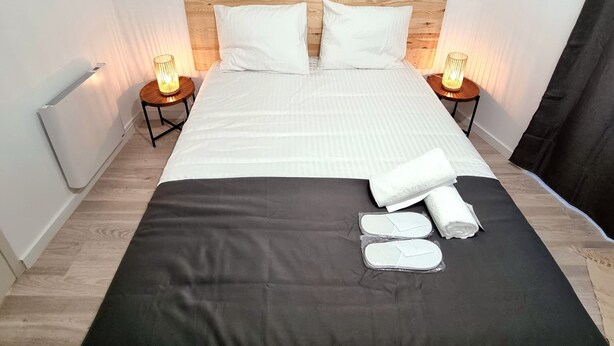Una cama bien hecha con colcha blanco y negro, dos almohadas blancas y una toalla doblada sobre la cama. Hay dos lámparas de noche con bombillas, un adorno en la pared sobre la cama, y una fotografía enmarcada en la pared.