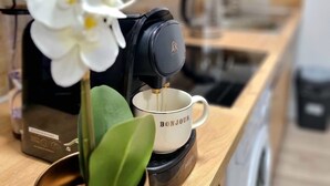 Una cafetera sobre una encimera de madera, con una taza de café blanca que dice "BONJOUR" y una orquídea en maceta.