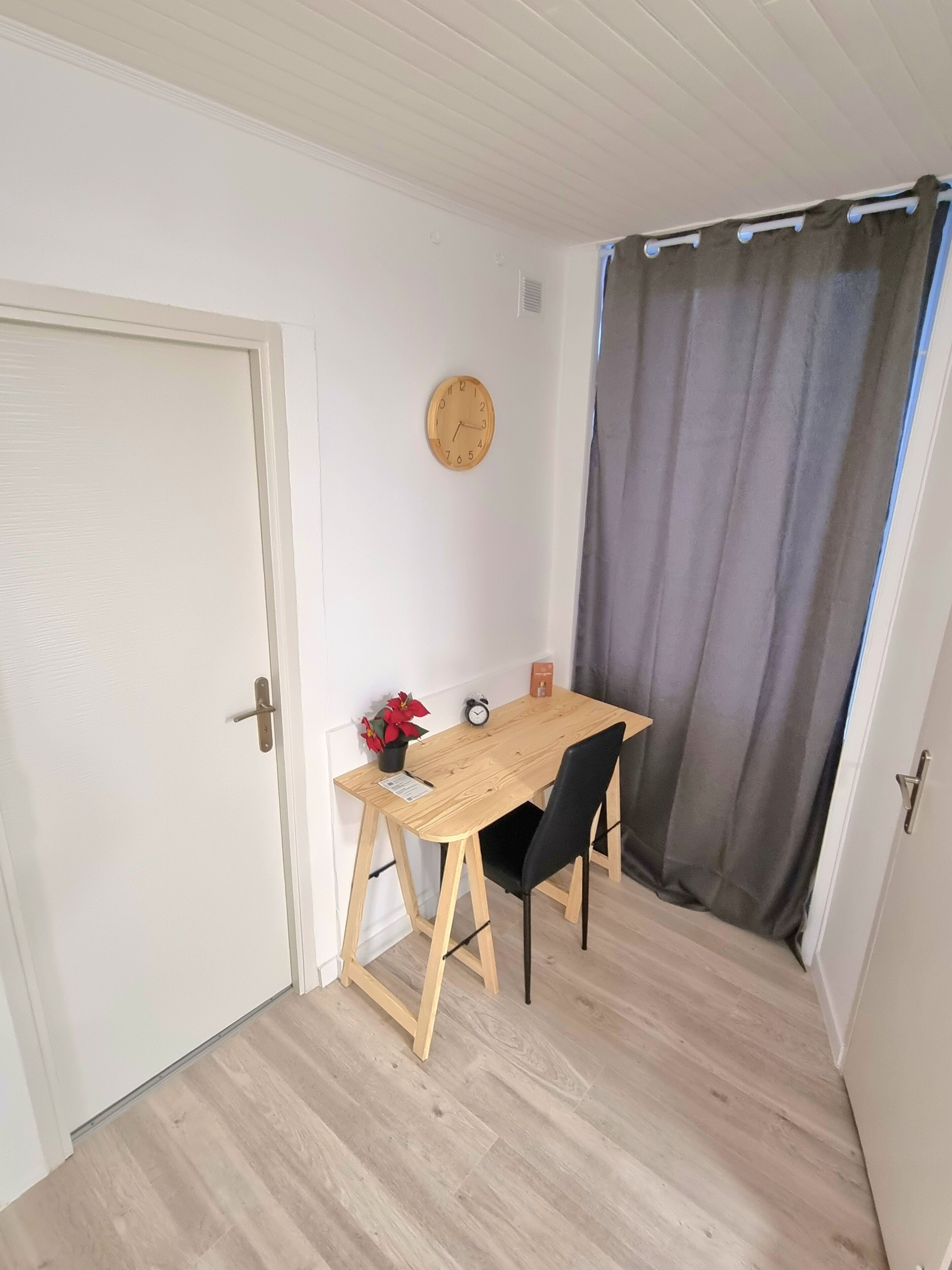 Et lille værelse med et træbord, to sorte stole, et ur på væggen, en dør og et gardin.