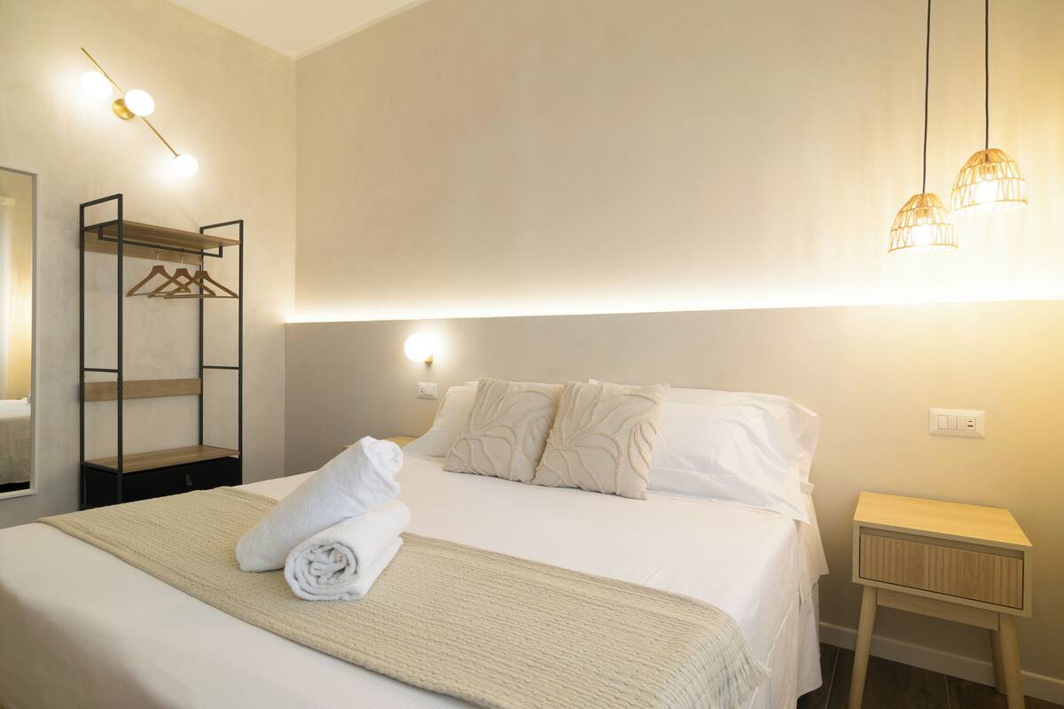 A neatly made bed with a rolled towel, a bedside table, a wall-mounted light fixture, and a hanging light.