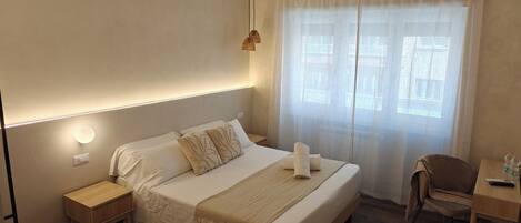 Habitación de hotel ordenada con cama, mesita de noche, silla y escritorio con otra silla.