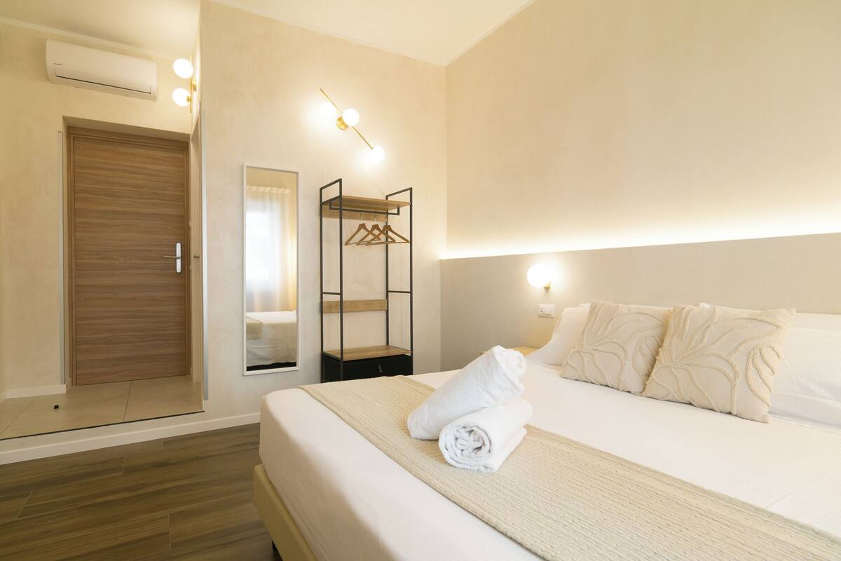 A hotel room with a wooden door, a mirror, a hanging coat rack, a neatly made bed with a rolled towel, and a bedside table.
