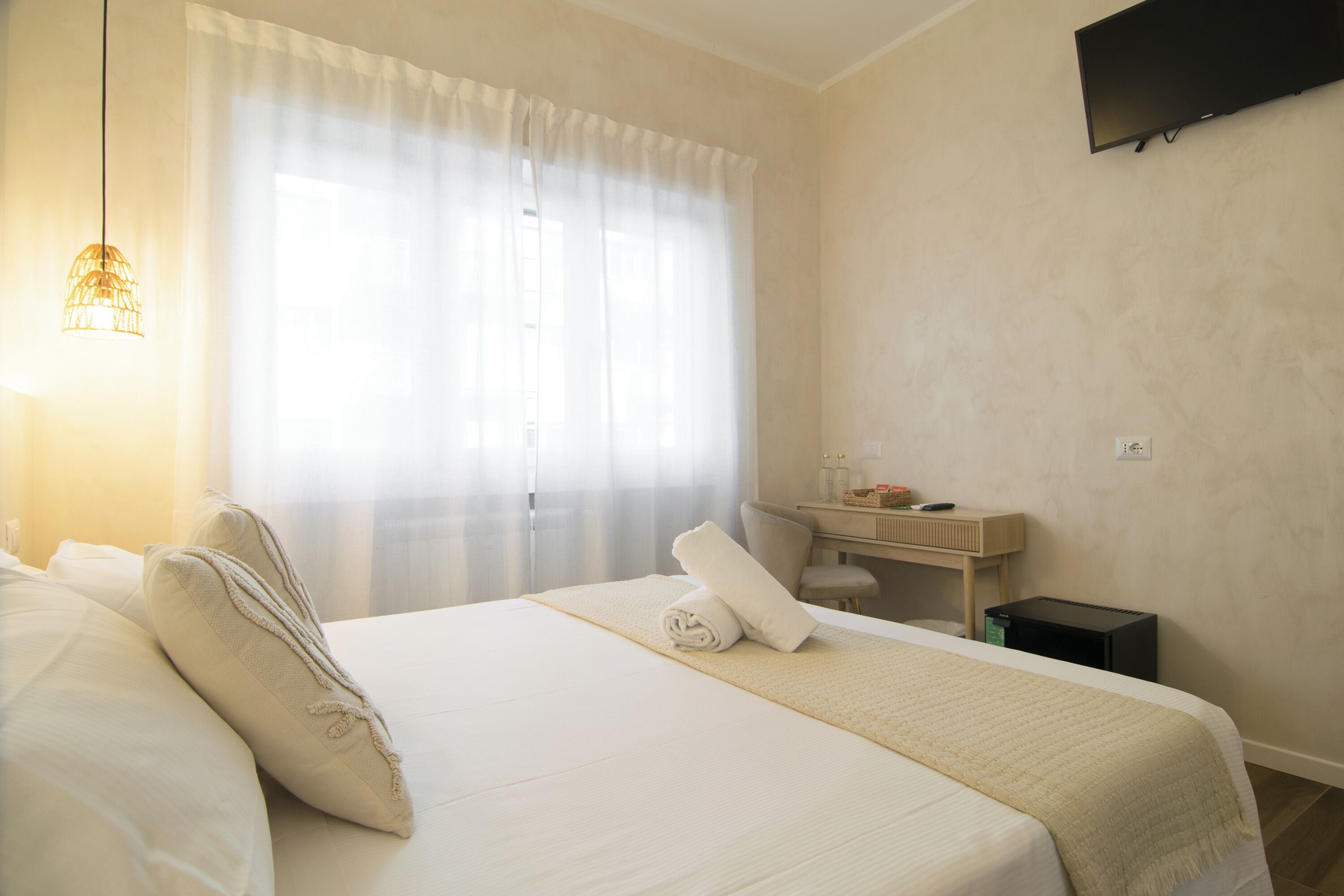 A hotel room with a bed, a bedside table, a TV mounted on the wall, and a window with curtains.