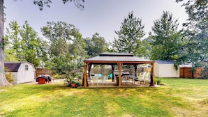 Un espace extérieur abrité, avec une table et des chaises, entouré d’une pelouse et d’arbres.