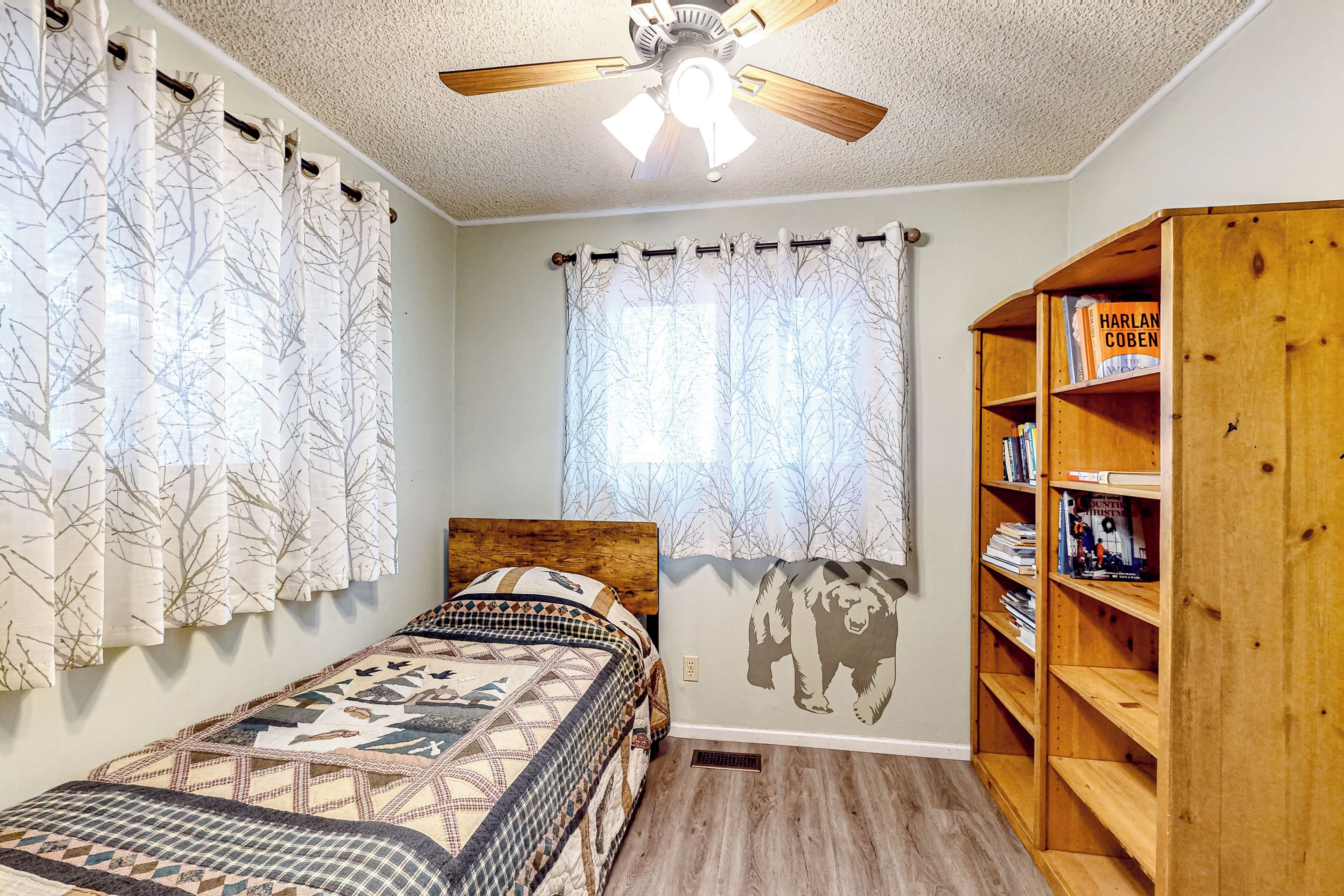 A bedroom with a bed, a ceiling fan, a window with curtains, and a bookshelf.
