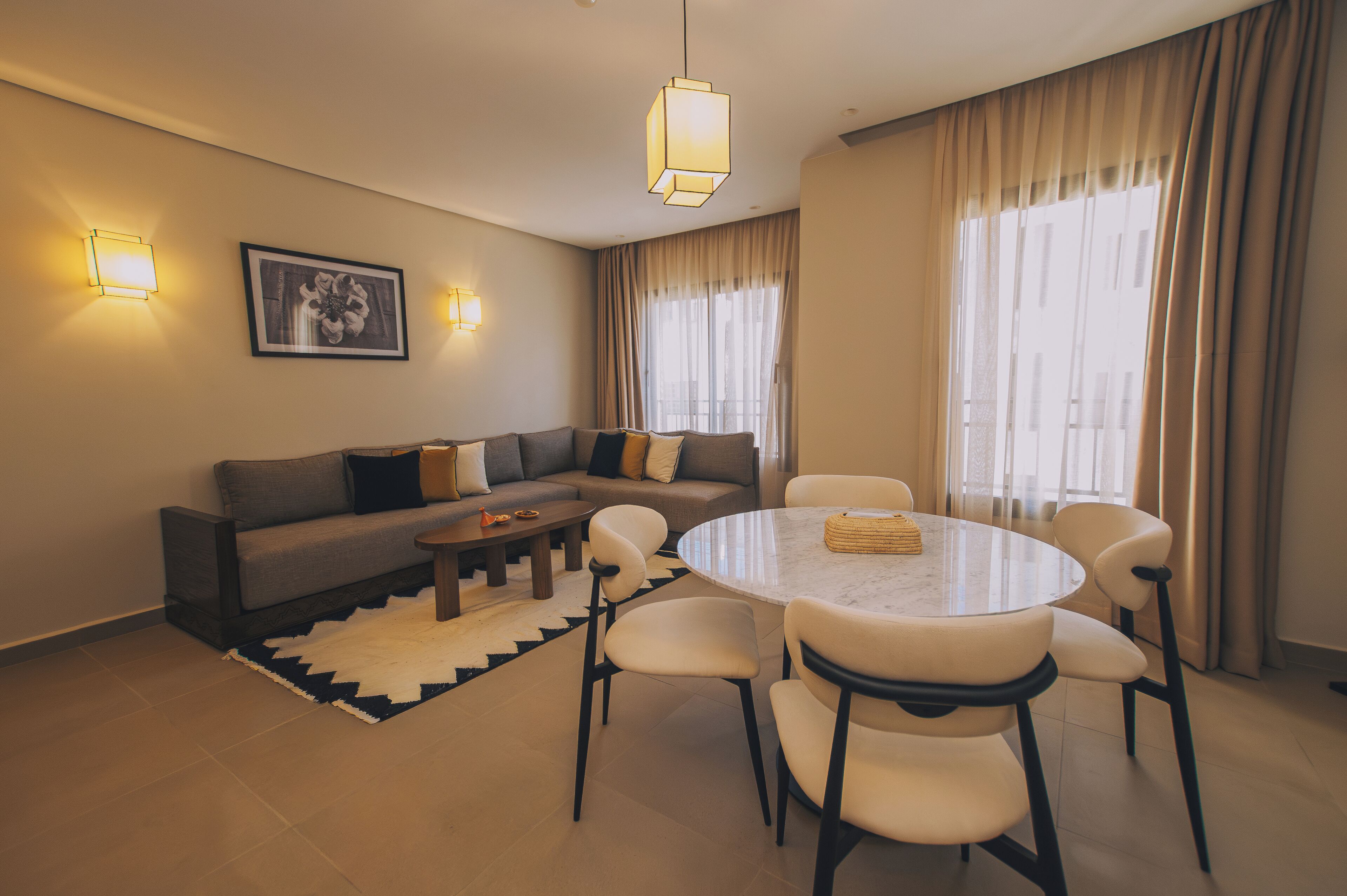 A modern living room with a sofa, a round dining table, and chairs. There are wall sconces and a large window with curtains.