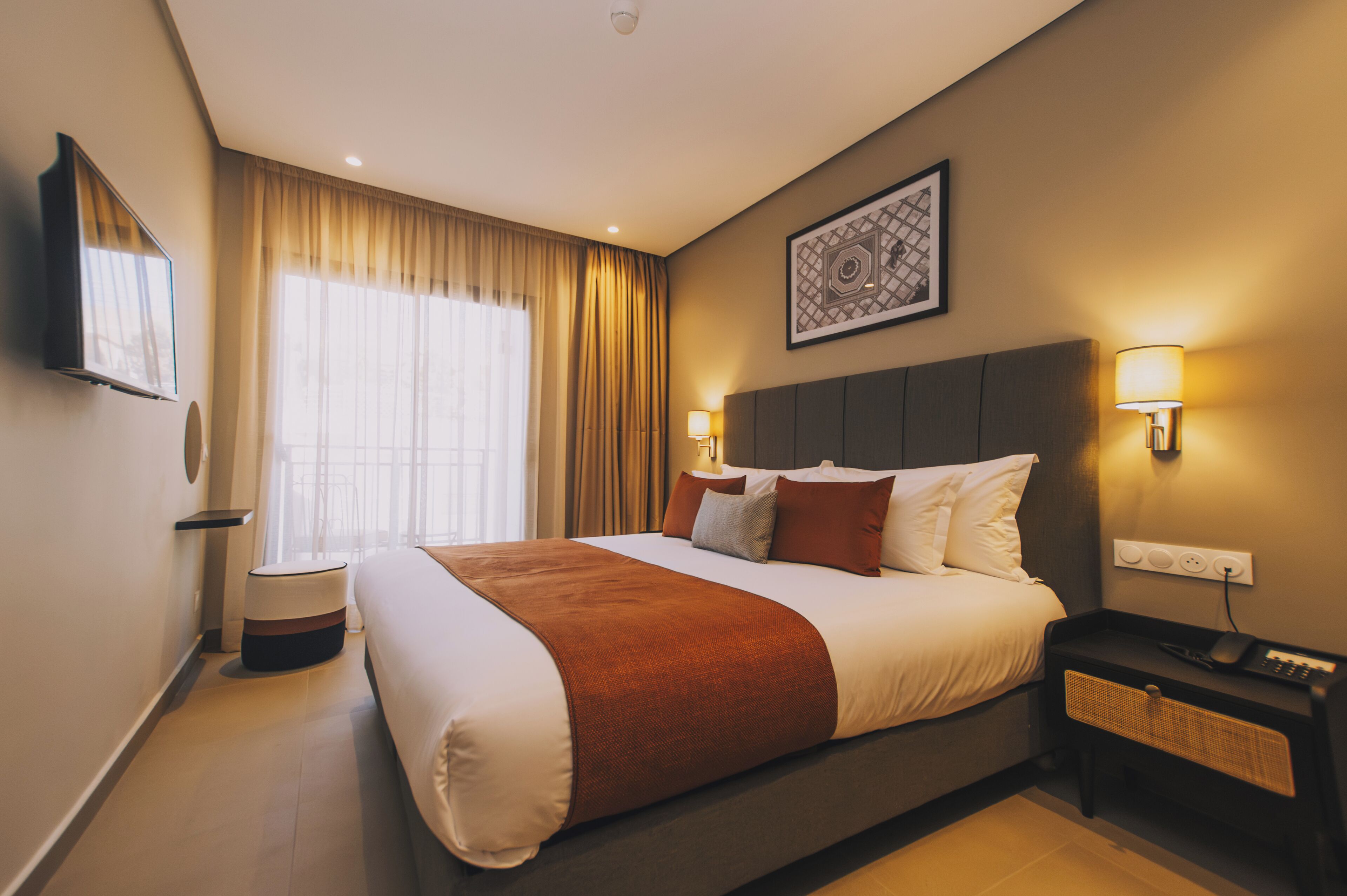 A hotel room with a large bed, a flat-screen TV, a bedside table with a lamp, and a window with curtains.