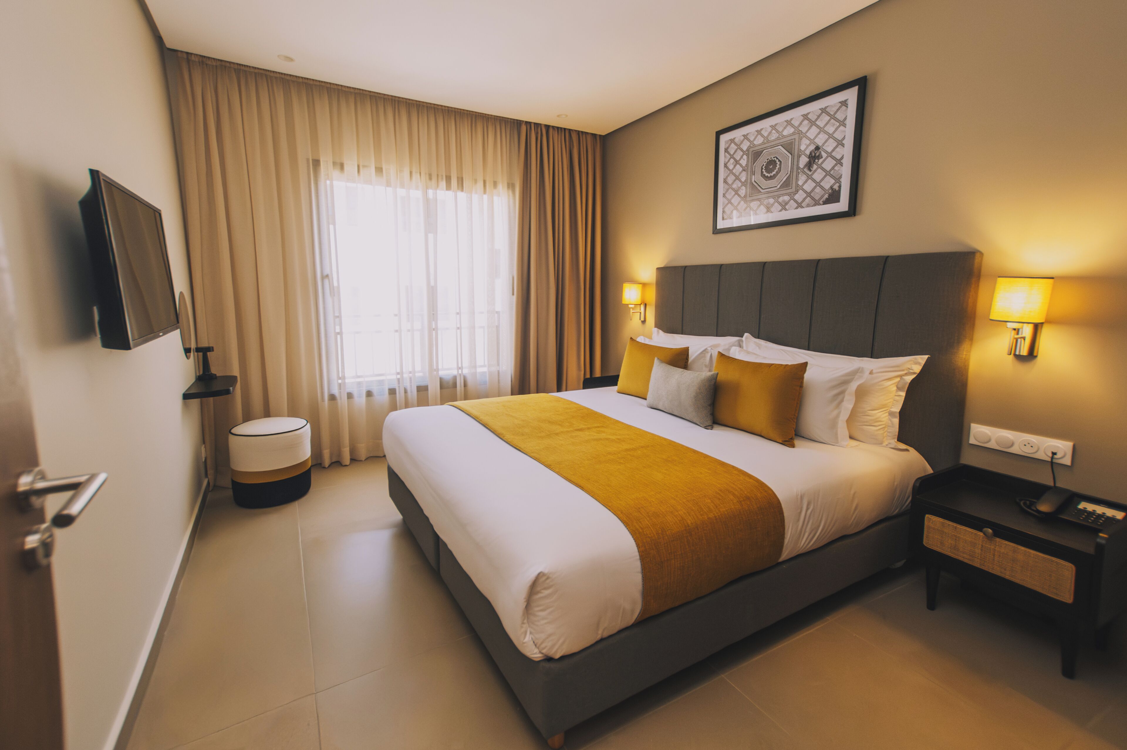 Habitación de hotel con una cama grande, una mesita de noche con lámpara, un televisor de pantalla plana en la pared y una ventana con cortinas.