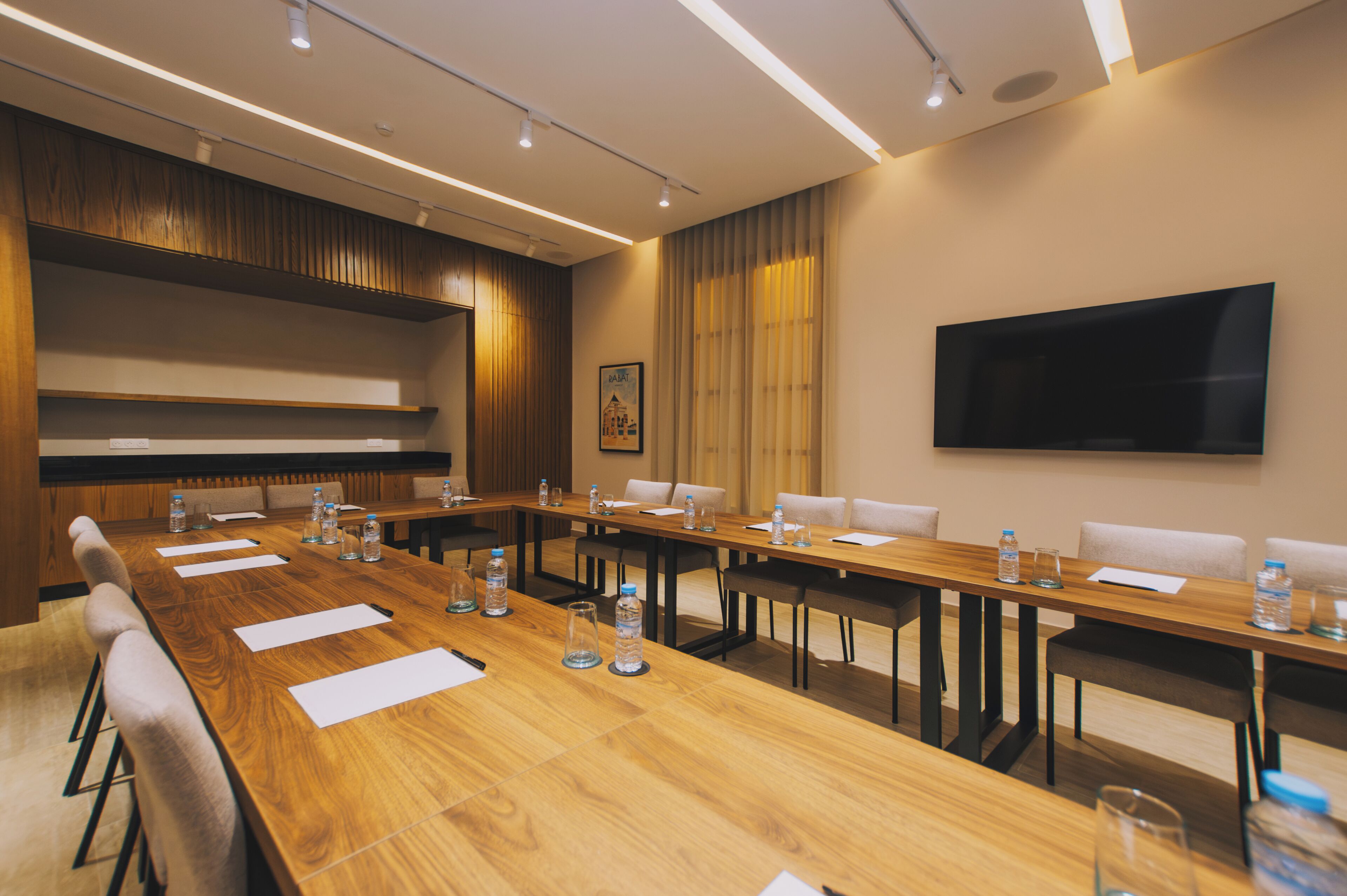 Une salle de conférence avec une longue table en bois, de nombreuses chaises, des bouteilles d’eau et un téléviseur à écran plat fixé au mur.