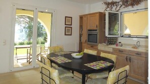 A dining area with a round table, yellow chairs, and a bowl of fruit. There is a kitchen with wooden cabinets and a window. - Villa Pelagos & guest house, undoubtedly has one of the best views in Corfu. (Corfu)