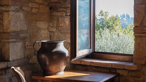 Een kamer met stenen muren, een houten tafel, een stoel en een groot raam met uitzicht op een bergachtig landschap.