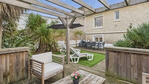 Een terras buiten met houten meubilair, een kleine tafel met bloemen en uitzicht op een stenen gebouw op de achtergrond.