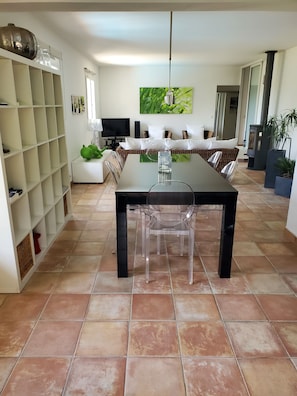Een moderne eethoek met een tafel met glazen blad, transparante stoelen, een wit kastje en een flatscreen televisie.