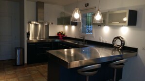 A modern kitchen with stainless steel appliances, a central island with bar stools, pendant lighting, and a window with a view. - Magnificent villa with private pool in a quiet location (vidauban)
